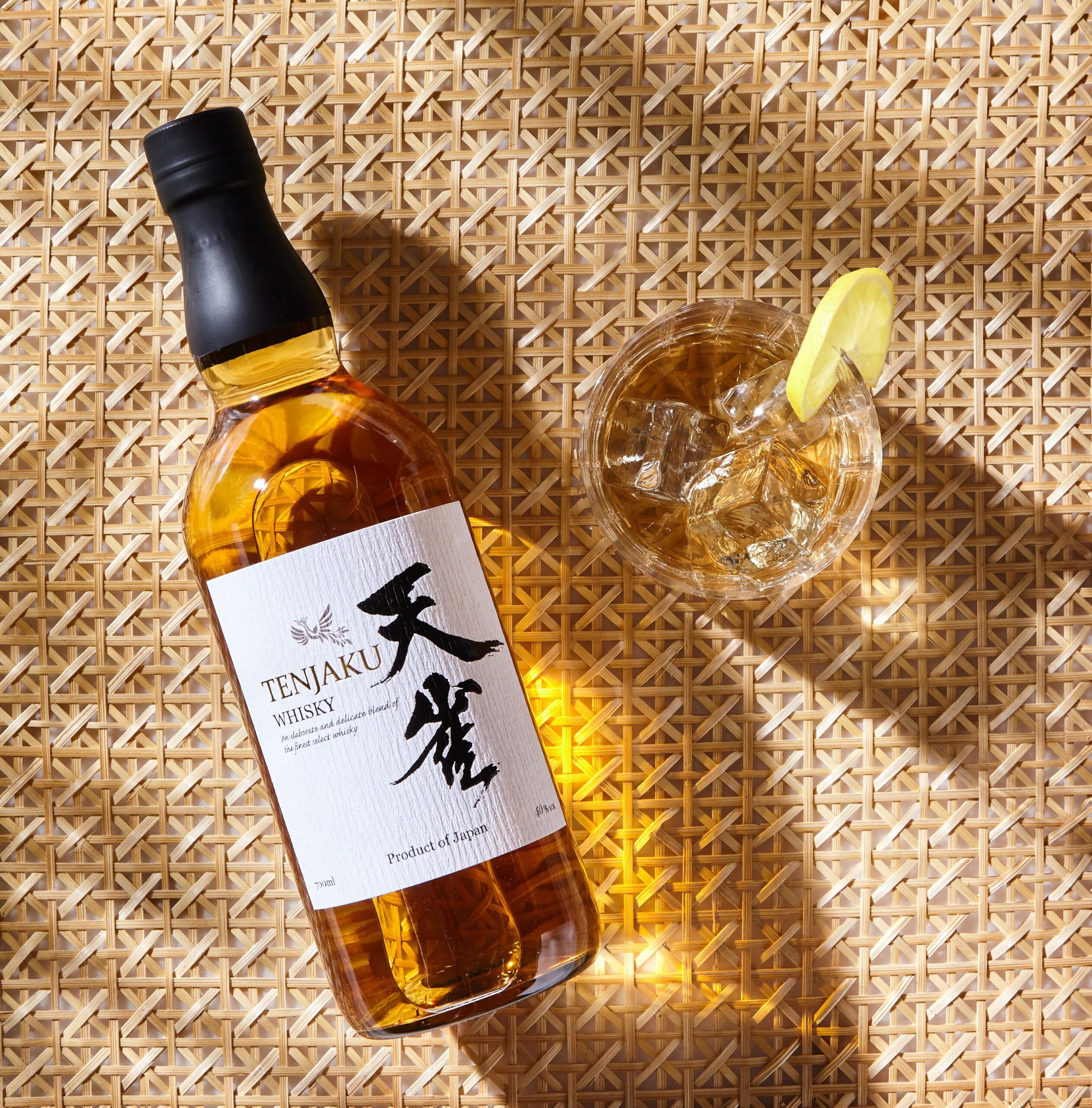 Tenjaku Whisky bottle beside an iced drink with lemon on woven matting.